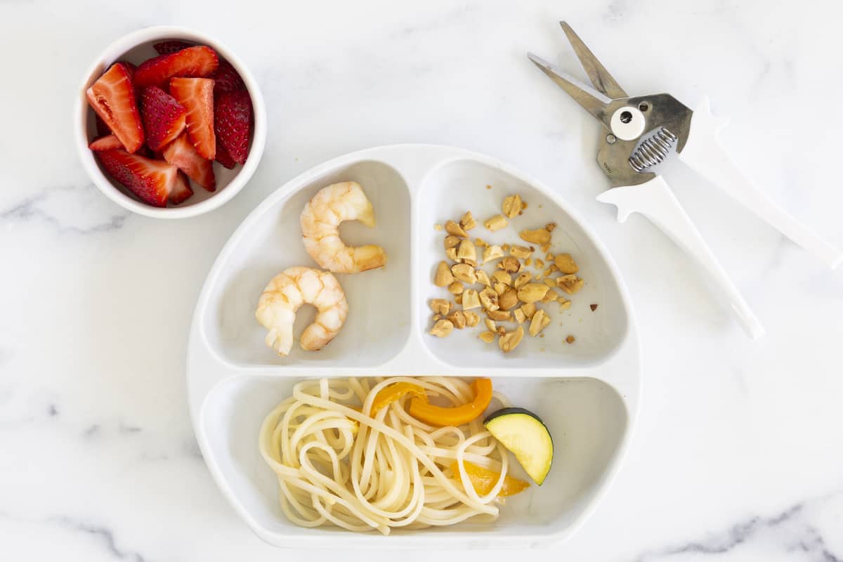 child's plate with shrimp and noodles with strawberries and scissors on side