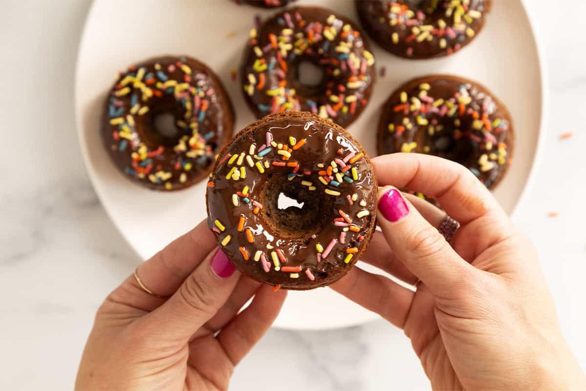 Hands holding chocolate baked donuts with frosting and sprinkles.