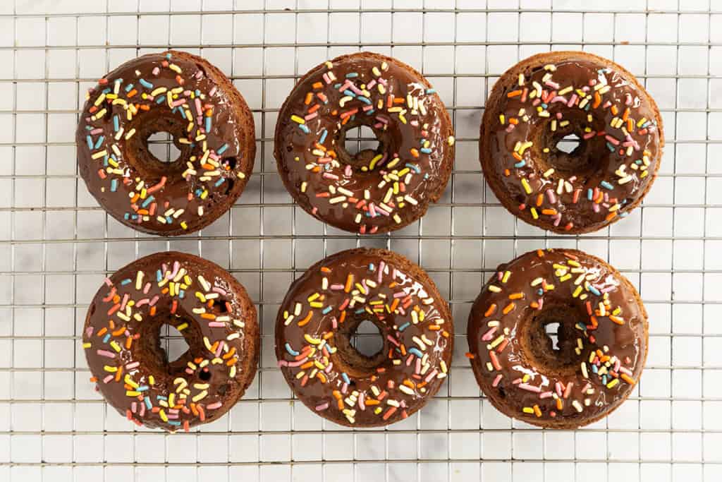 Chocolate baked donuts with frosting and sprinkles on cooling rack.