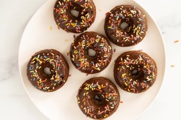 Chocolate baked donuts with frosting and sprinkles on white plate.