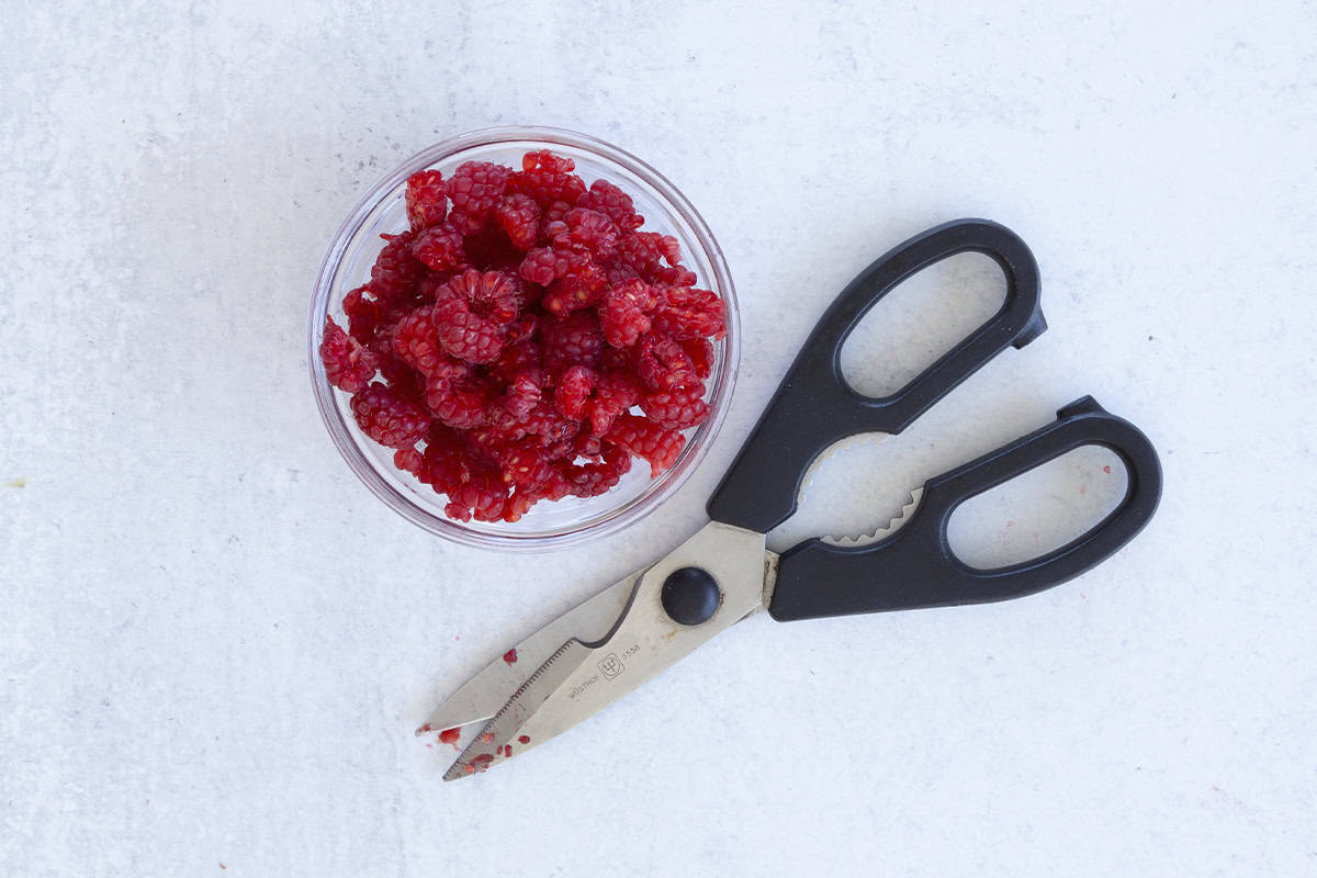 Raspberries being cut with scissors into glass bowl.