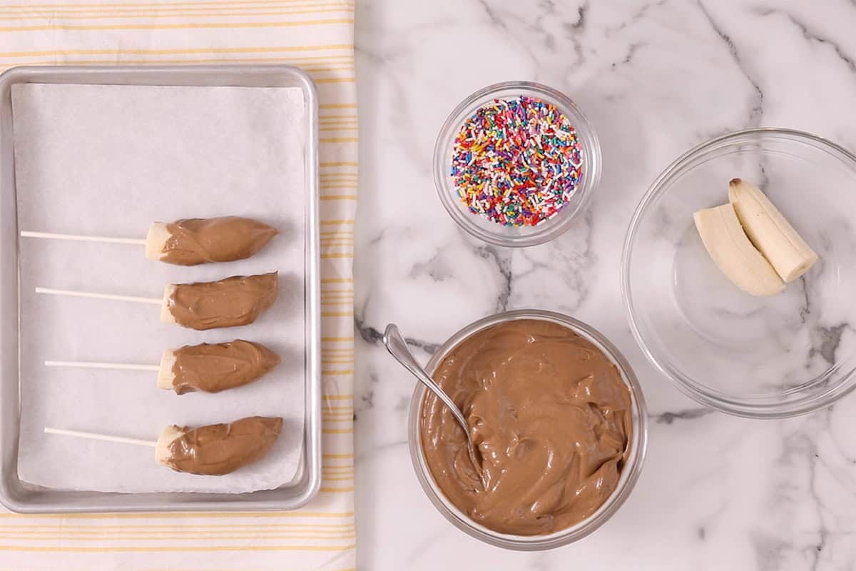 Chocolate covered bananas on cooking sheet with sprinkles on side.