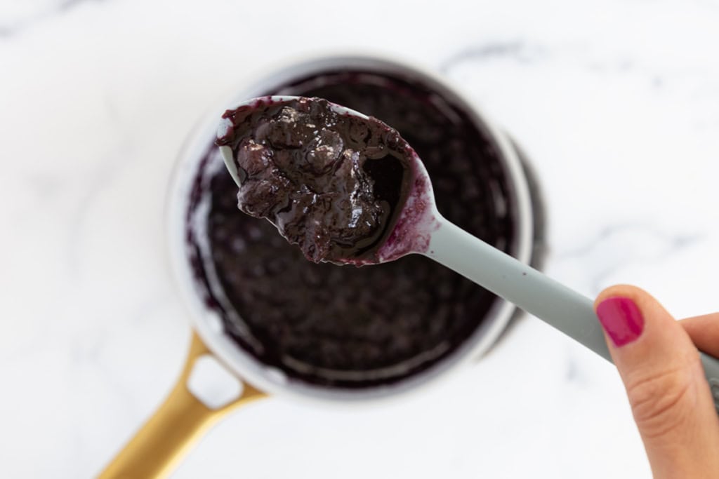 Pan with cooked blueberry sauce and hand holding spoon.