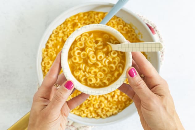 Hands holding bowl of pasta in broth with pan underneath.