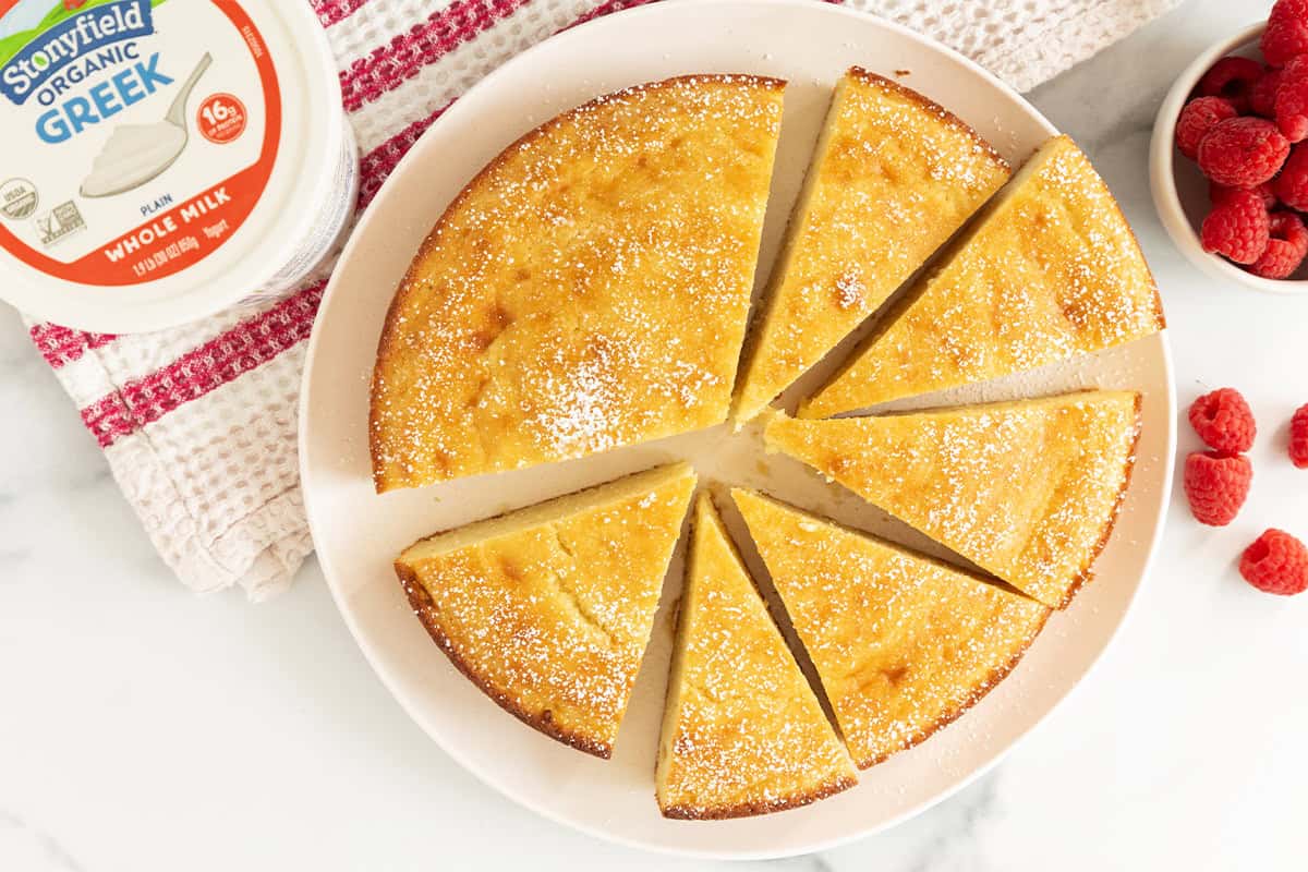 Greek yogurt cake on white plate cut into slices with yogurt container on side. 