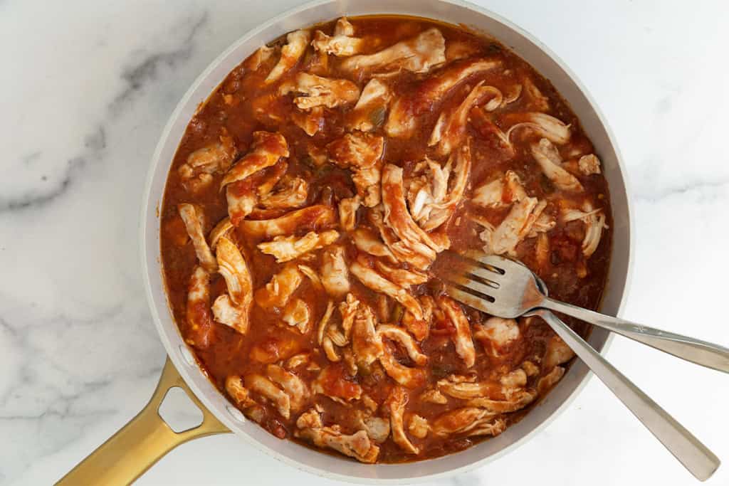 Chicken being shredded with forks in frying pan.