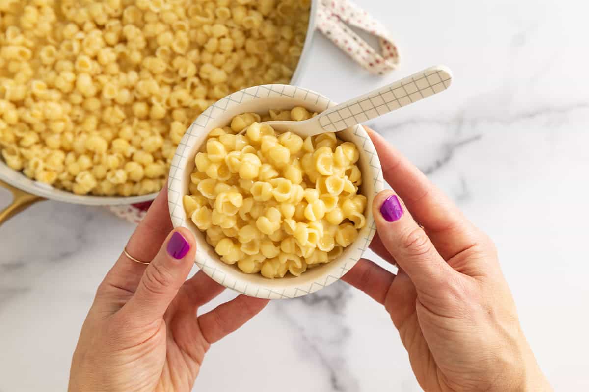 Hands holding bowl of mac and cheese over pan. 