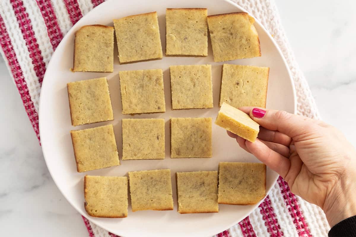 Banana blondies cut into squares on white plate with hand holding square.