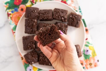 Chocolate snack cake cut into squares on plate with hand holding slice.